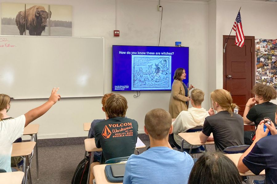 Marie Stango stands at the front of a high school classroom. Students in the class focus their attention toward her. One student is raising his hand.